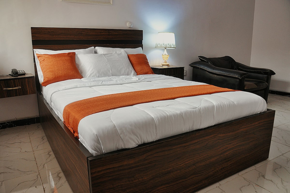 A neatly made bed with a wooden headboard, a bedside lamp, and a chair in a hotel room.