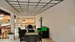 Ein überdachter Außenbereich mit Tisch und Stühlen, einem grünen Sofa und Blumentöpfen.