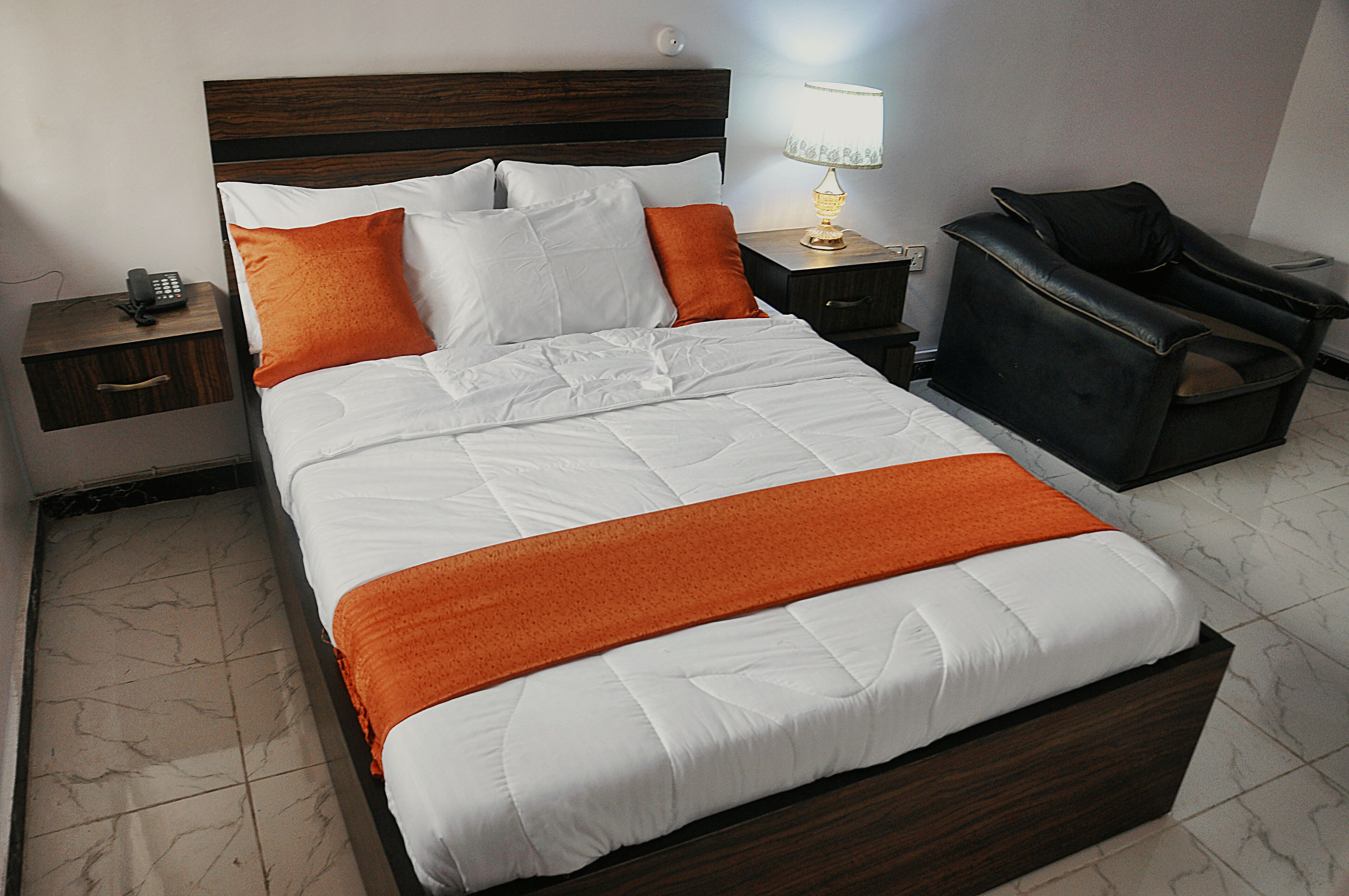 A hotel room with a large bed, a nightstand with a lamp, a chair, and a telephone.