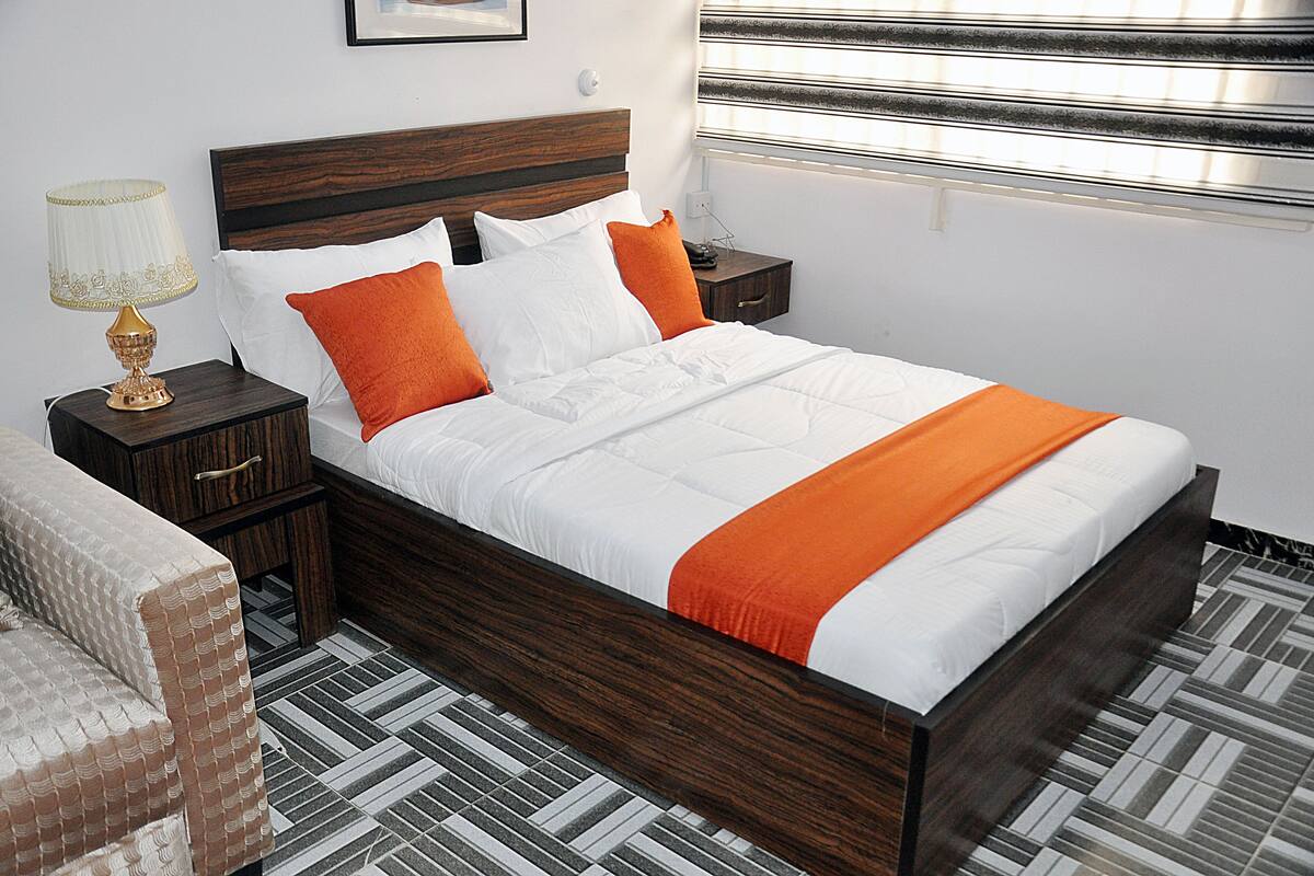 A neatly made bed with white and orange bedding, a wooden headboard, and a nightstand with a lamp.