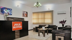 Ein Empfangsbereich mit einem Schild, das "JOLIE SUITES" zeigt, einem schwarzen Empfangstresen, Ledersofas, einer Kristallleuchter und einem großen Fenster mit Jalousien.
