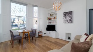 Ein modernes Wohnzimmer mit Holzboden, einem Essbereich mit Tisch und Stühlen, einem Bücherregal, einem Flachbildfernseher und einem Kamin.