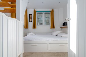 A bedroom with a bed, white walls, a window with yellow curtains, a TV on a shelf, and a wooden ceiling structure.