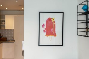 A framed abstract painting on a white wall in a modern kitchen with white cabinets and a grey backsplash.