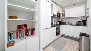 A modern kitchen with white cabinets, a stainless steel range hood, and a built-in oven. There is a bookshelf with books and a red sign, and a small rug on the floor. - GuestReady - Amazing spot near Gilbert Park (London)