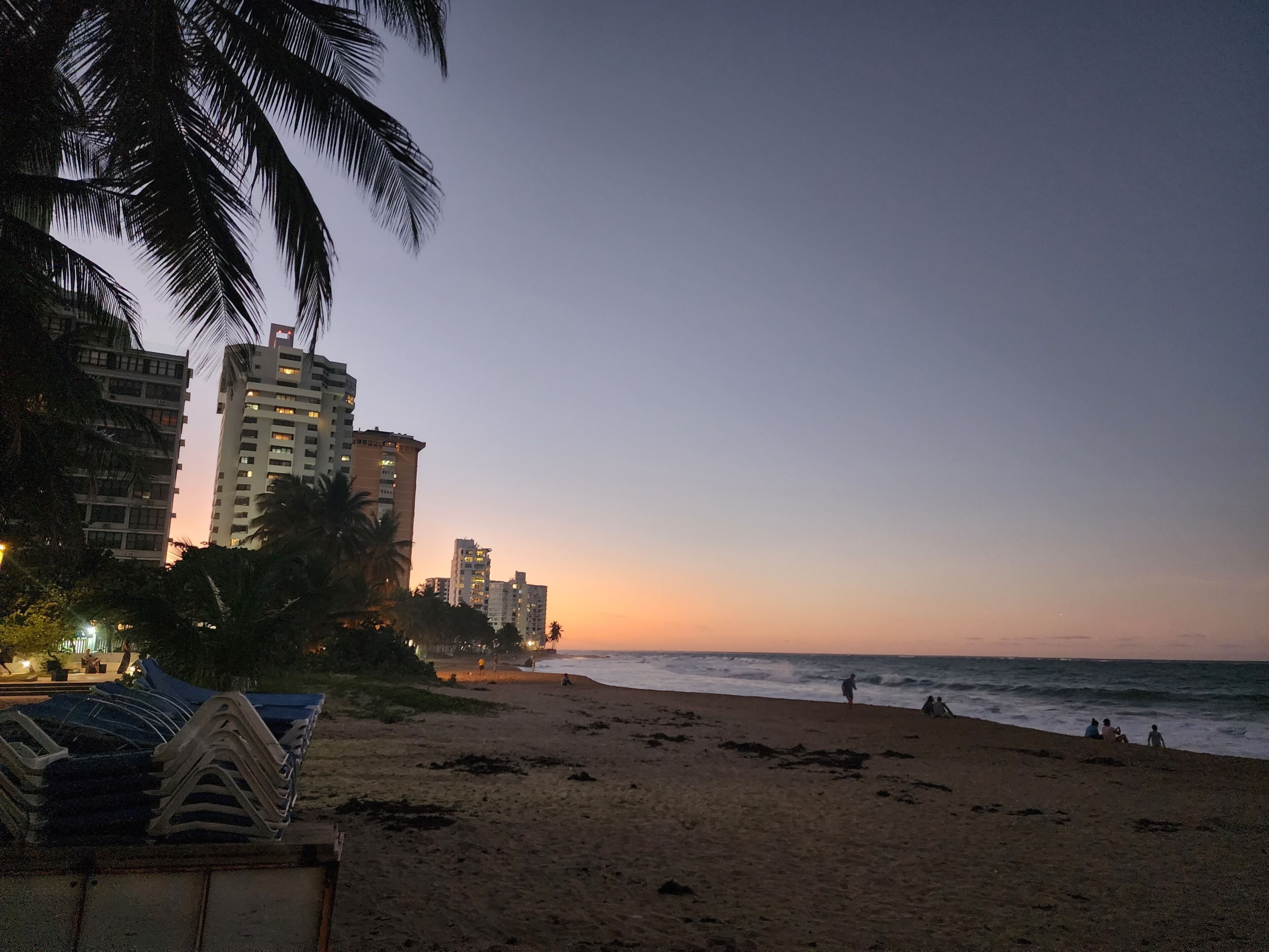 A beach with a row of tall buildings in the background, a palm tree on the left, and a stack of plastic chairs in the foreground.