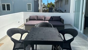Een balkon buiten met een tafel en stoelen, een bank met kussens en uitzicht op omliggende gebouwen.