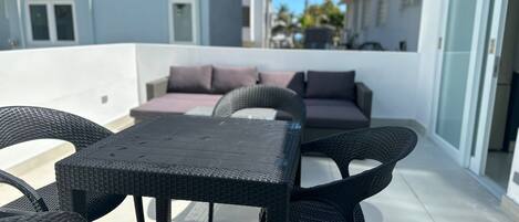 A rooftop terrace with a black woven table and chairs, a sofa with cushions, and a view of modern buildings in the background.
