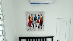 Een net opgemaakt bed met witte lakens en een zwart hoofdeinde, een schilderij van abstracte figuren aan de muur, en een witte deur aan de rechterkant.