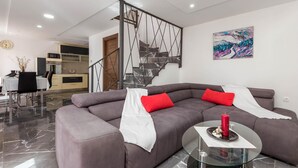 A modern living room with a grey sofa, red cushions, a glass coffee table, and a staircase with a metal railing. - 3 bedroom lovely home in Jurdani (Jurdani)