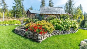 Uma cabana rústica com base de pedra e telhado de madeira, cercada por um jardim bem cuidado com diversas plantas e flores.