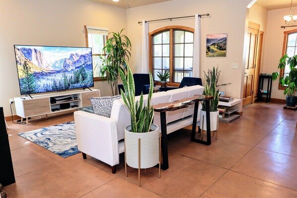 A modern living room with a flat-screen TV, white sofas, a coffee table, and potted plants.