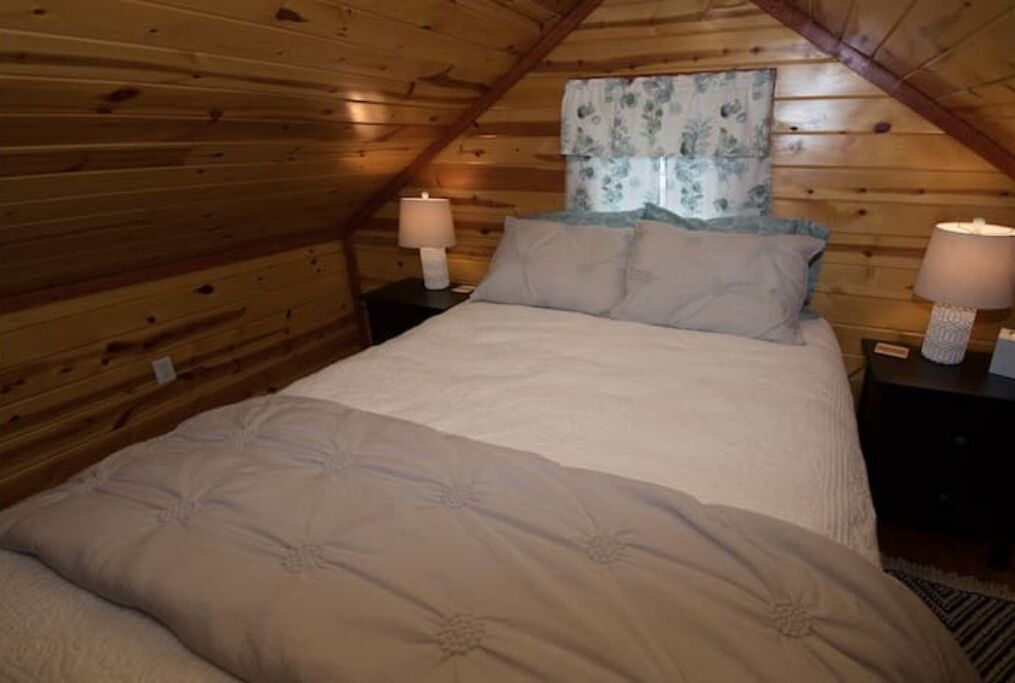 A cozy bedroom with a wooden ceiling, a bed with a beige duvet, two bedside tables with lamps, and a floral curtain.