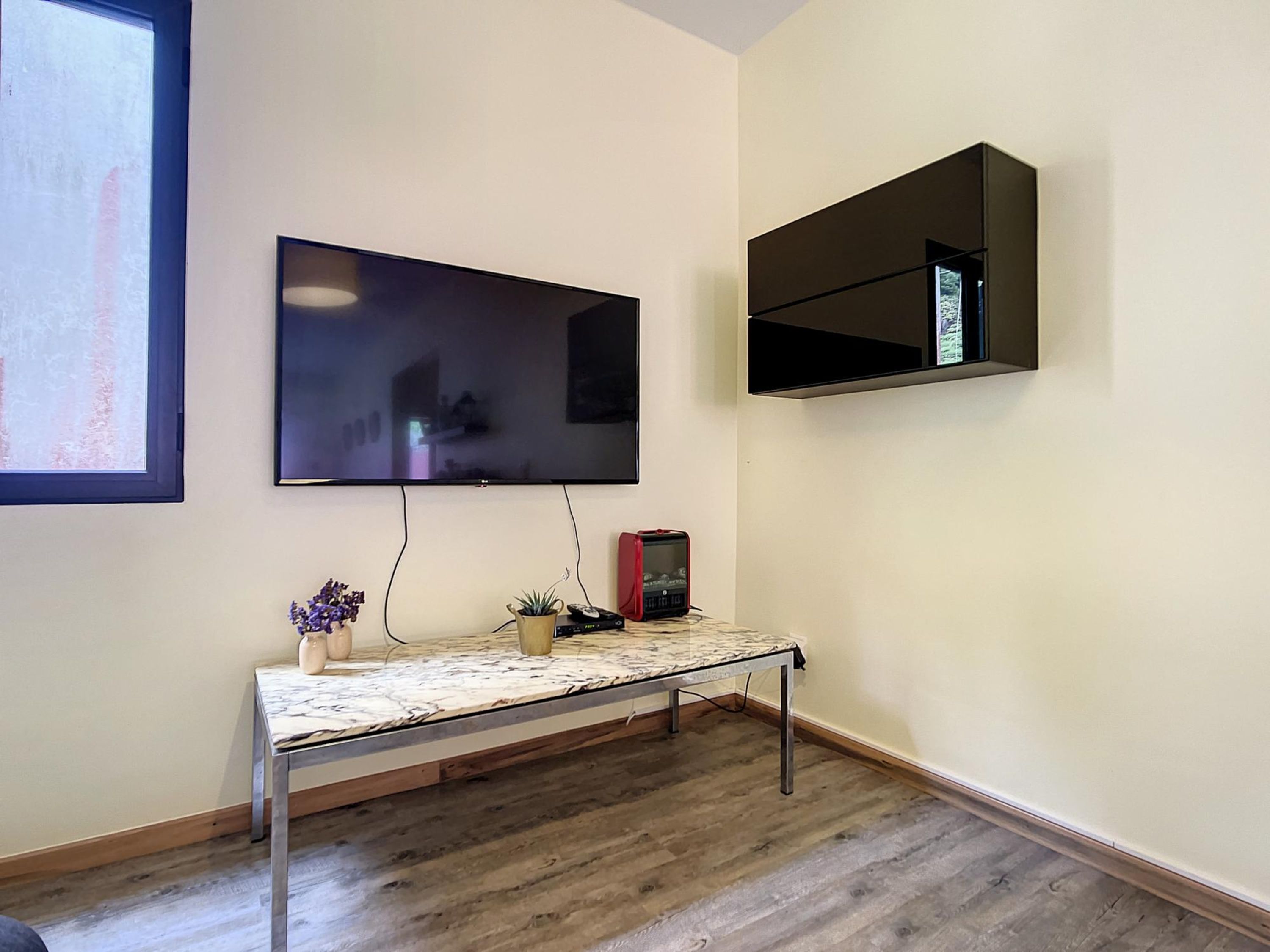 A room with a flat-screen TV mounted on the wall, a small table with a vase and a plant, and a shelf above the table.