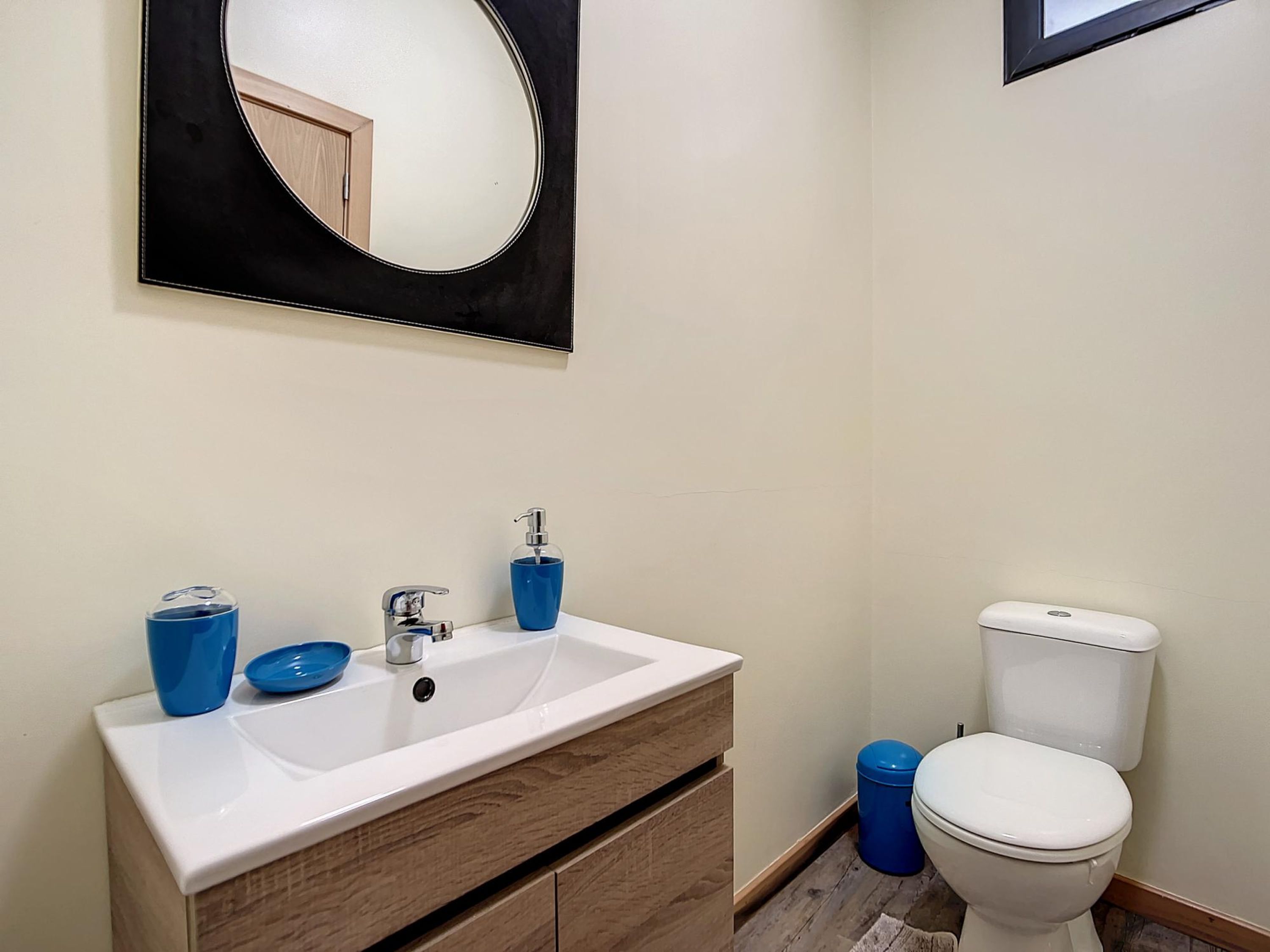 Une salle de bain avec un lavabo blanc, un miroir, des toilettes et une poubelle bleue.