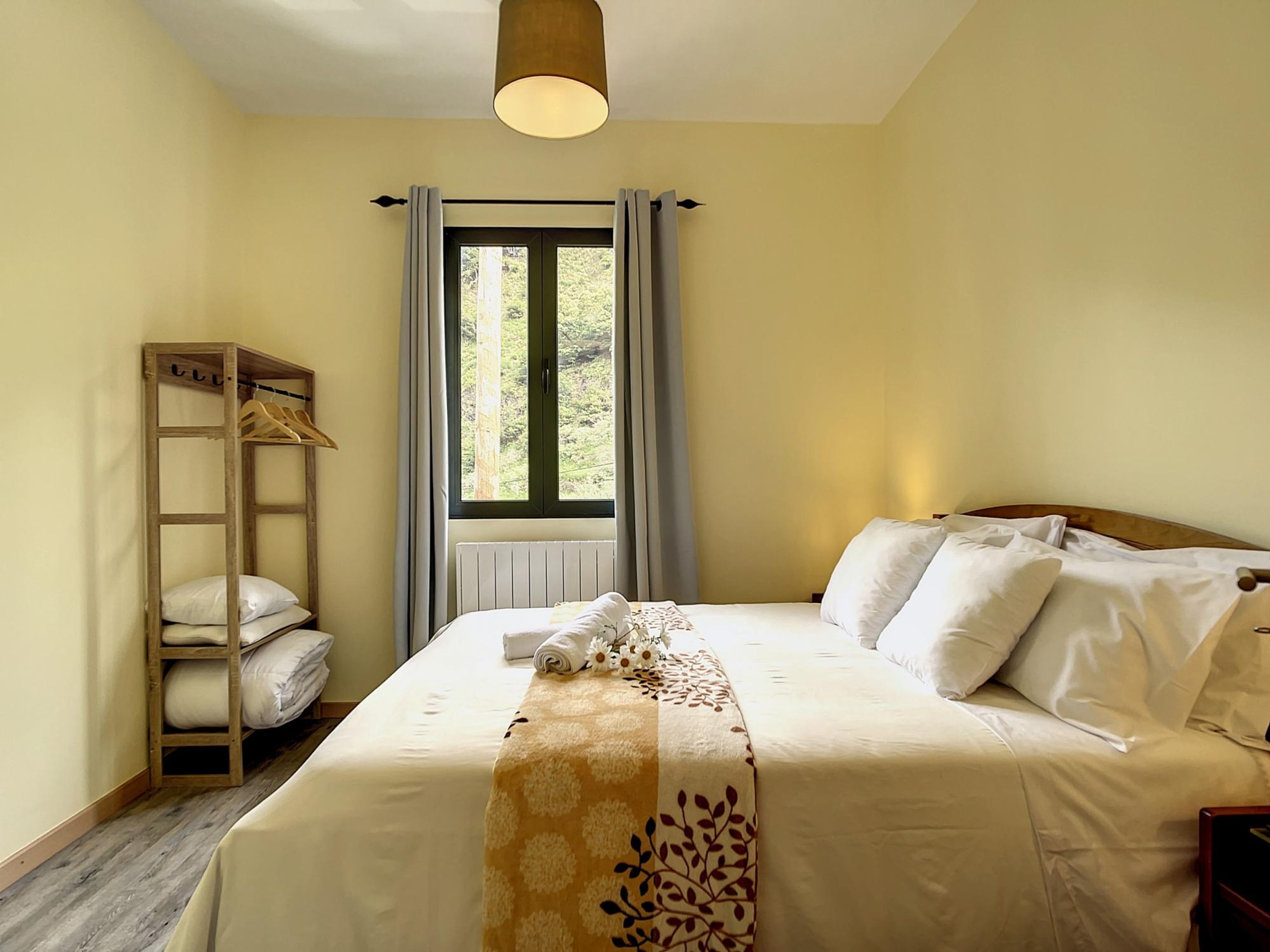 A bedroom with a bed, a wooden shelf with hanging clothes, a window with curtains, and a radiator.