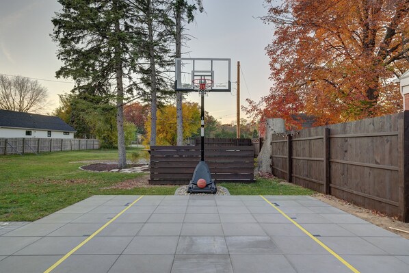 A basketball court with a hoop, surrounded by a wooden fence and trees with autumn foliage.
