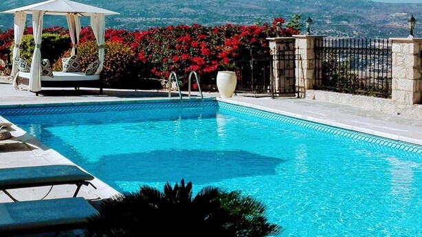 Ein klares, blaues Schwimmbecken mit einer Laube, Liegestühlen und Blick auf rote Blumen und Hügel im Hintergrund.