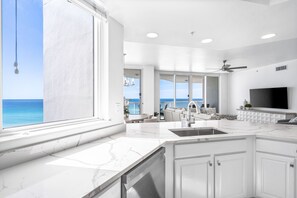 A modern kitchen with a large window overlooking the ocean, a built-in dishwasher, and a flat-screen TV mounted above the sink. - Stunning, fully renovated 5 Star Beachfront Condo (Destin)