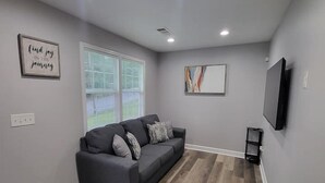Una sala moderna con un sofá gris, piso de madera, un televisor de pantalla plana y una ventana con persianas.