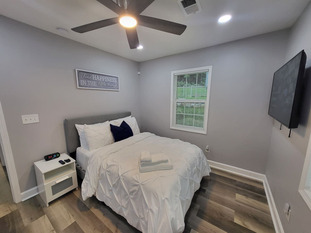 A bedroom with a bed, nightstand, ceiling fan, wall-mounted TV, and a window with blinds.