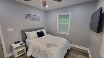 A bedroom with a bed, nightstand, ceiling fan, wall-mounted TV, and a window with blinds.