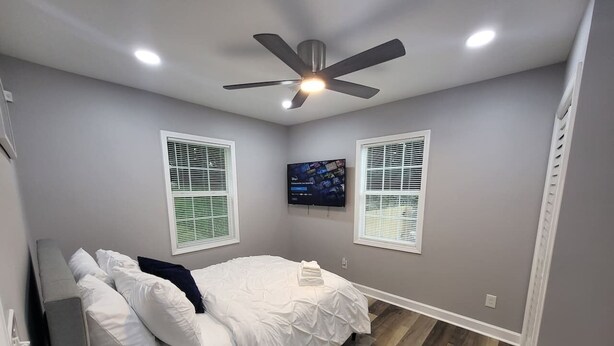 Un dormitorio con cama, ventilador de techo, televisor montado en la pared y dos ventanas con persianas.