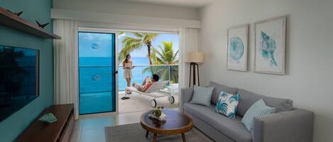 A modern living room with a grey sofa, a wooden coffee table, and a TV on a wooden stand. There is a balcony with a view of the ocean and palm trees.
