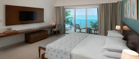 A modern hotel room with a large bed, a flat-screen TV, a desk, and a balcony with a view of the ocean.