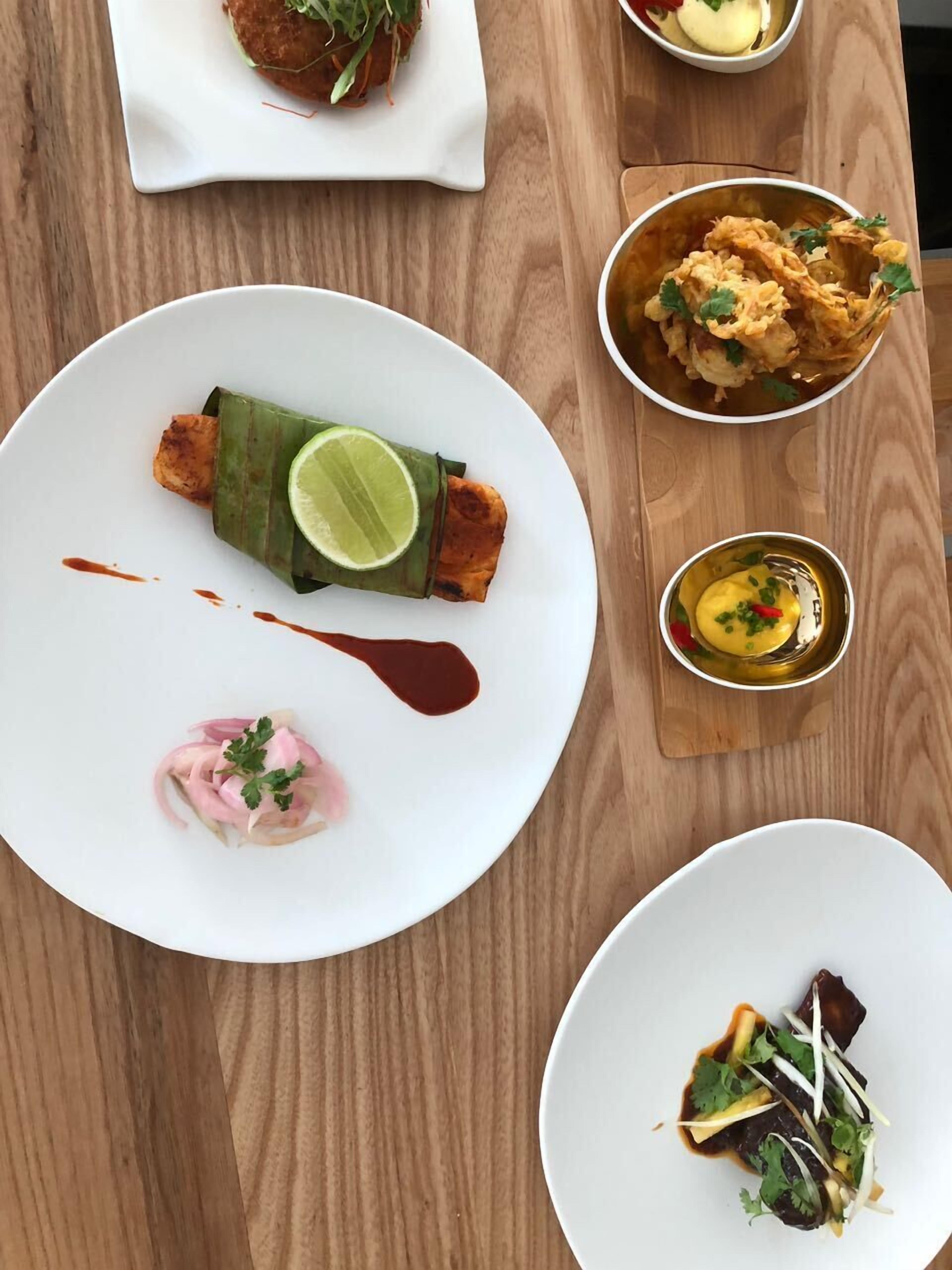 Uma mesa de madeira com diversos pratos, incluindo um prato com uma fatia de limão, uma tigela com alimentos fritos e um pequeno recipiente com molho amarelo.