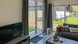 Un salon moderne doté d’un grand téléviseur, d’un canapé avec des coussins, d’une table basse agrémentée d’objets décoratifs et d’une porte-fenêtre coulissante donnant sur un jardin.