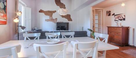 A dining area with a white table and chairs, a kitchen with a TV, and a wooden ceiling.