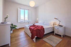 Une chambre à coucher avec un lit, des tables de chevet, une commode, une lampe, une fenêtre avec des volets, une plante en pot et une vue sur la ville.