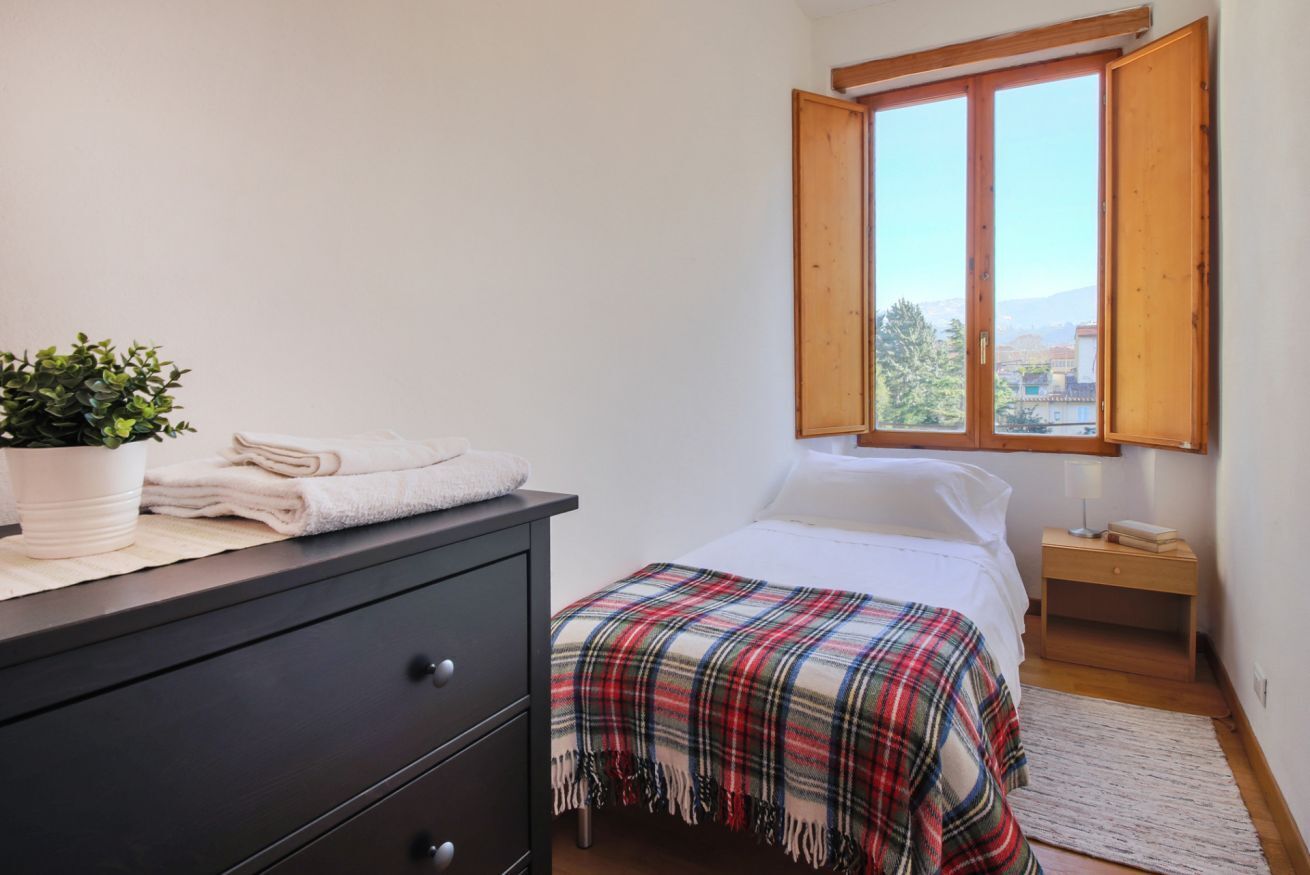 A bedroom with a bed, a nightstand, a dresser, a window with an open shutter, and a potted plant.