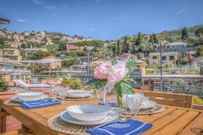 A dining table set with plates, glasses, and a vase of flowers, overlooking a hillside with buildings. - CASA SOLE - Terrace with sea view, relaxation and WiFi (Camogli)