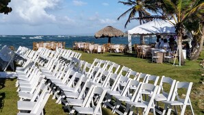 A beachside event setup with white plastic chairs arranged in rows, a thatched umbrella, and a tent with people inside. - Villas Rossen  (Puerto Plata)