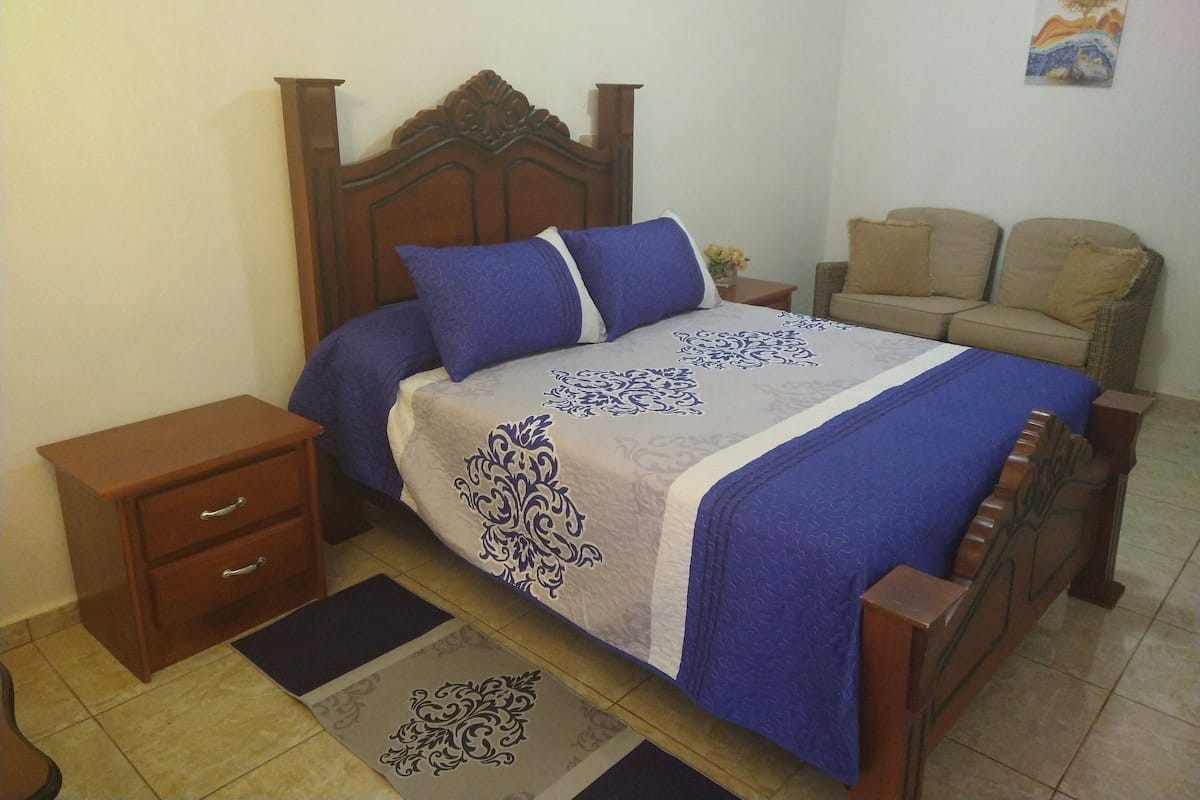 A neatly made bed with a wooden headboard, a nightstand with drawers, a chair, and a small table with a vase of flowers.