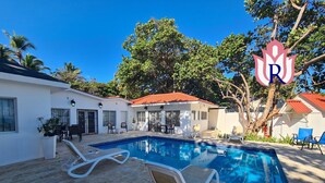 A swimming pool with lounge chairs, a patio area, and a white building with a red roof. - Villas Rossen  (Puerto Plata)