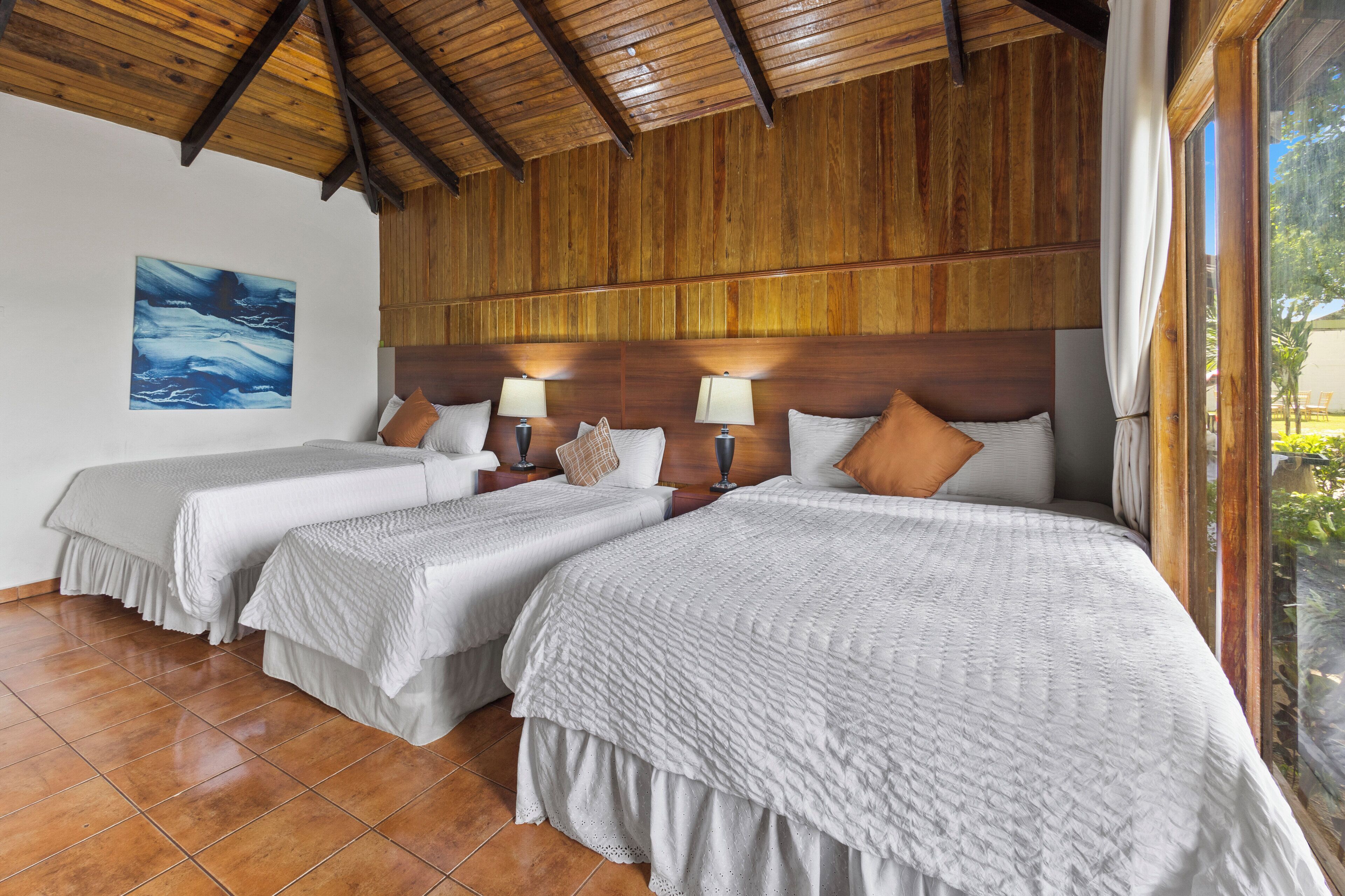 A hotel room with two beds, wooden ceiling beams, a large window, and a painting on the wall.