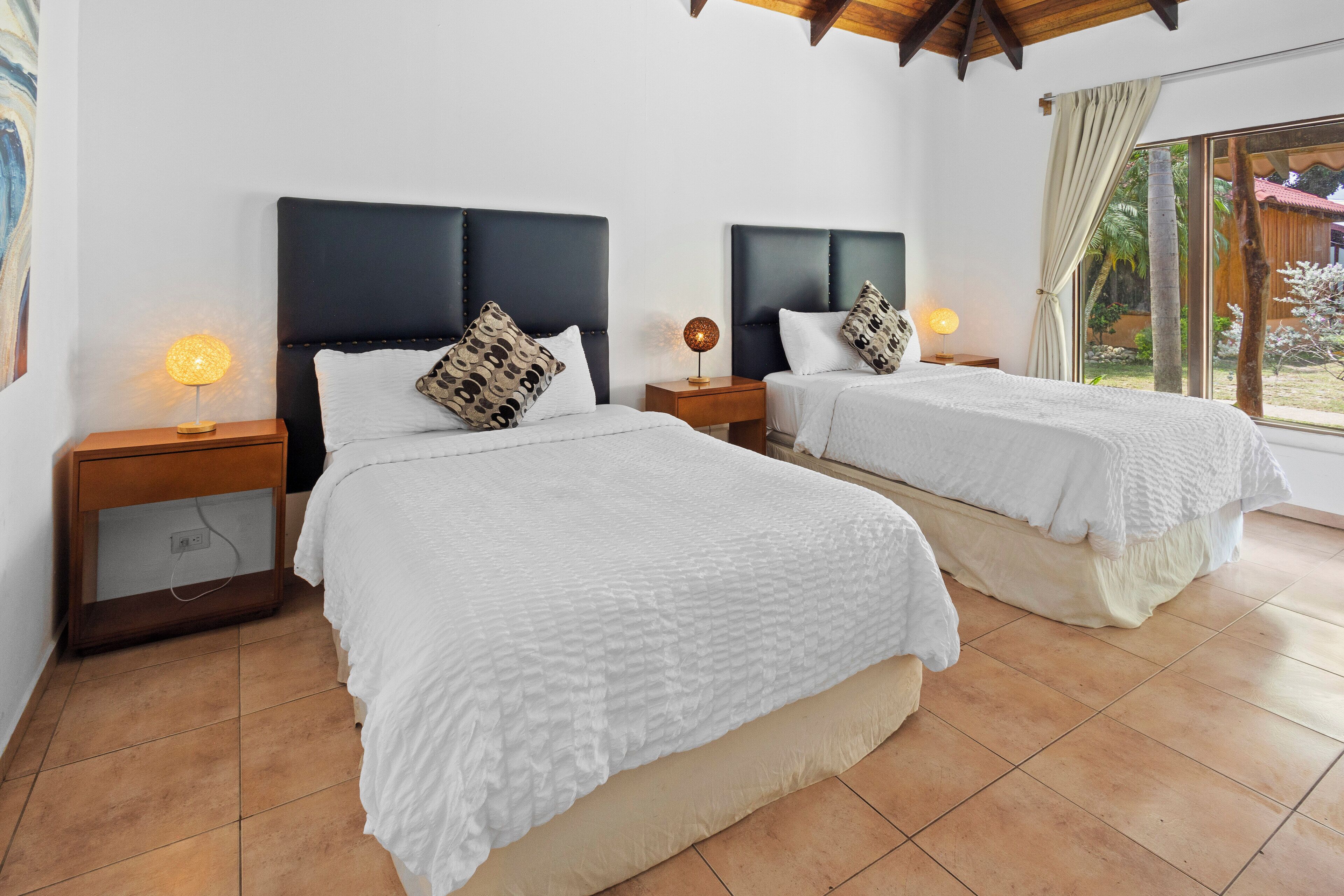 A bedroom with two beds, each with a headboard, bedside lamps, and a window with a view of the outdoors.