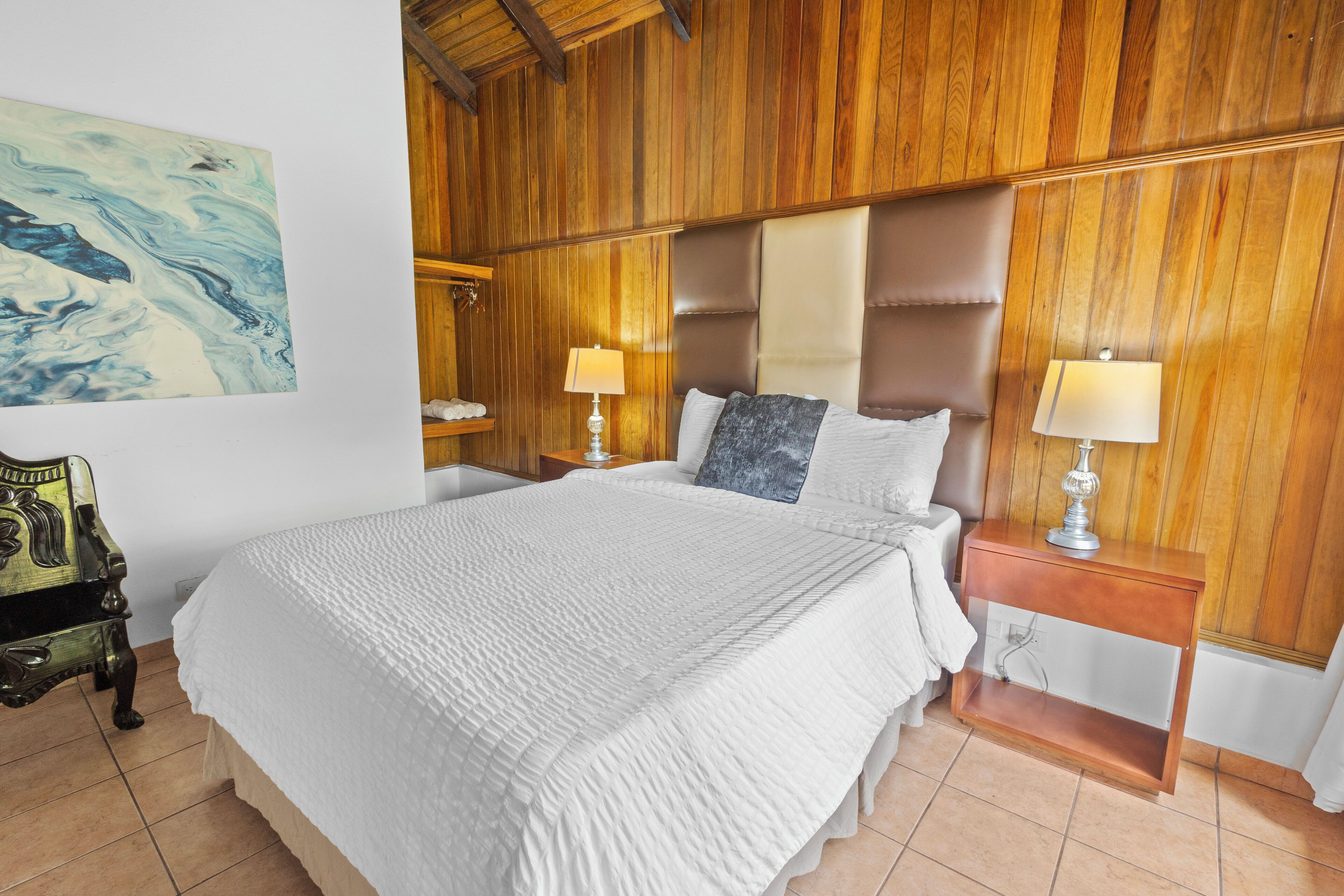Un dormitorio con una cama grande, paredes de paneles de madera, dos mesitas de noche con lámparas y un banco con lámpara.