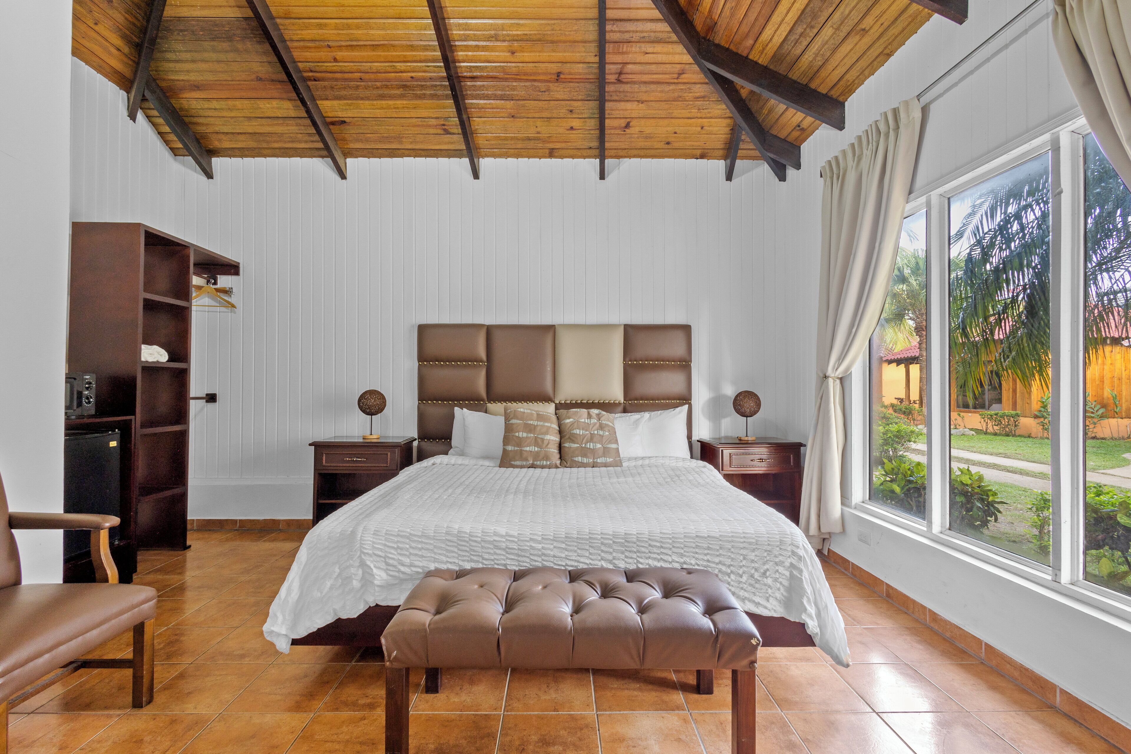 Un dormitorio con una cama grande, un banco de madera, un armario y una ventana con vistas a zonas verdes.