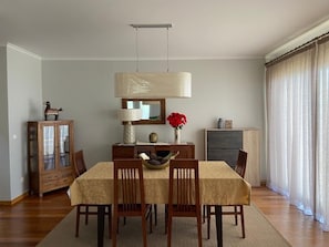 A dining area with a table set for four, a wooden cabinet, and a large window with curtains. - The Best Sea View in Madeira -Casa Farol  (Fajã da Ovelha)