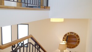 Una escalera con barandilla de madera y balaustradas negras que conduce a un segundo piso. Un espejo redondo cuelga de la pared sobre un aparador de madera con una lámpara y objetos decorativos.
