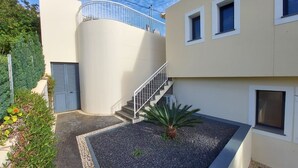 Un edificio moderno con una pared exterior curva, una escalera que lleva a un balcón y un pequeño jardín con grava y una palmera.