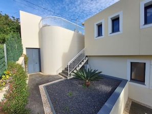 A modern building with a curved exterior wall, a staircase leading to a balcony, and a small garden area with gravel and a palm tree. - The Best Sea View in Madeira -Casa Farol  (Fajã da Ovelha)