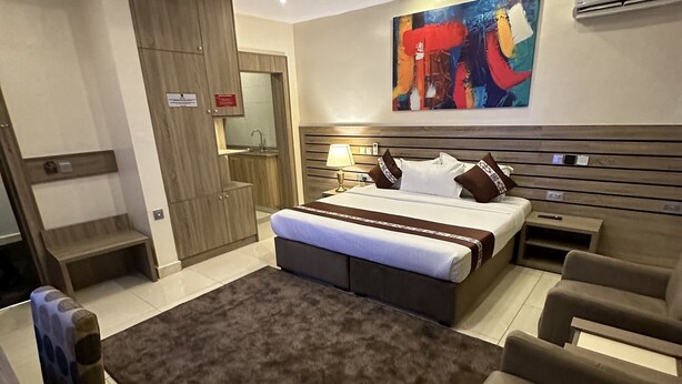 A modern bedroom with a large bed, a sitting area with sofas, a small table, and a colorful abstract painting on the wall. - Moz One Executive Guest Rooms-Tourggourt (Abuja)