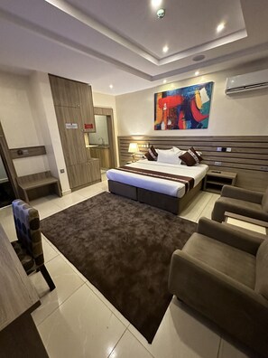 Ein modernes Schlafzimmer mit einem großen Bett, einer Sitzecke mit Sofas, einem kleinen Tisch und einem farbenfrohen, abstrakten Gemälde an der Wand.