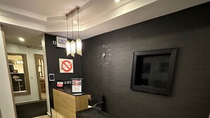 A modern reception area with a dark wall, a wooden counter, a black bench, and a mounted TV. - Moz One Executive Guest Rooms-Tourggourt (Abuja)