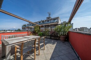 A rooftop terrace with a wooden table and chairs, potted plants, and a view of a modern building with a satellite dish. - GuestHost - Cozy apartment of 143 sqm, able to accommodate up to 5 people, located on the third floor of a building with lift in the heart of Udine. The location is great! In about 10 minutes walk you can reach Via Mercatovecchio, Piazza della Libertà a (Udine)
